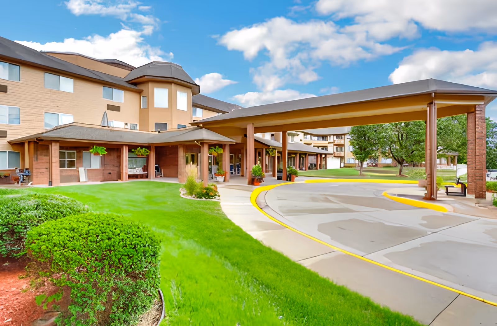 Exterior view of Holiday Country Squire facility showing a covered driveway entrance with a curved sidewalk, green manicured lawn, bushes, and trees under a partly cloudy blue sky.
