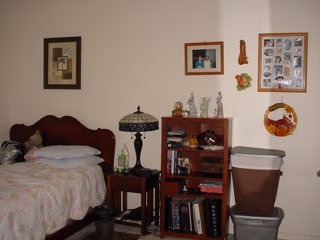 A small bedroom with a wooden bed and nightstand, Tiffany-style lamp, a bookshelf holding books and decorations, and stacked storage bins beneath framed photos on the wall.