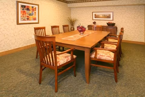 A dining room with a long wooden table surrounded by eight wooden chairs with cushioned seats. The table has a decorative runner and a centerpiece with flowers. The room has patterned wallpaper, carpeted floor, and framed artwork on the walls. A wooden sideboard with a bowl of fruit is against the far wall.