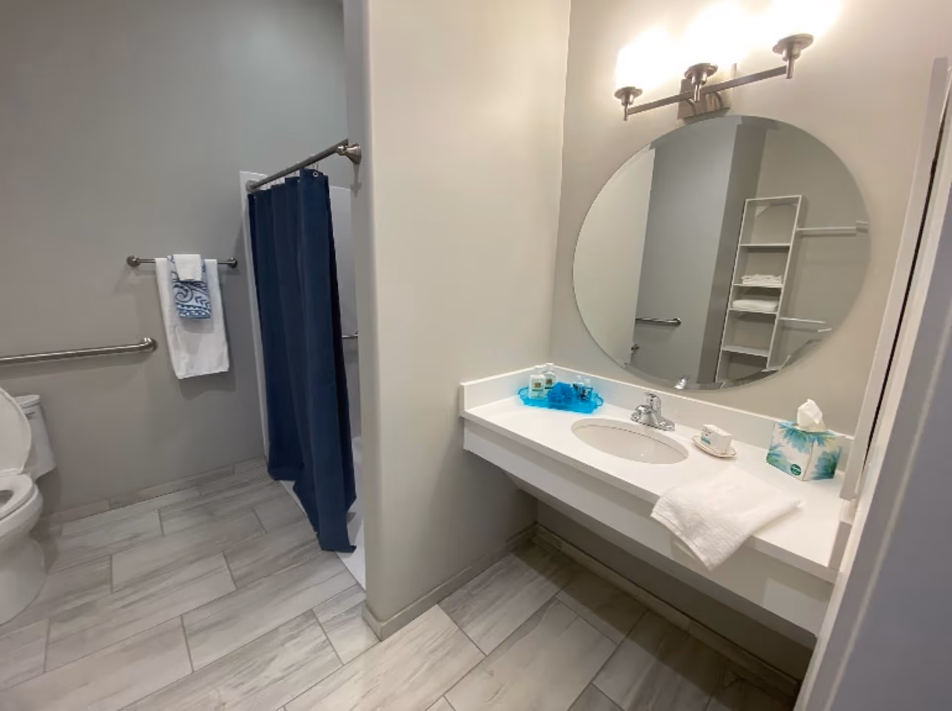 A clean and modern bathroom with light gray tiled floor, a white toilet, a shower with a dark blue curtain, a white towel rack with two towels, and a white vanity with a round mirror above it. The vanity has a sink, a tissue box, a soap dish, and some toiletries on a blue tray.