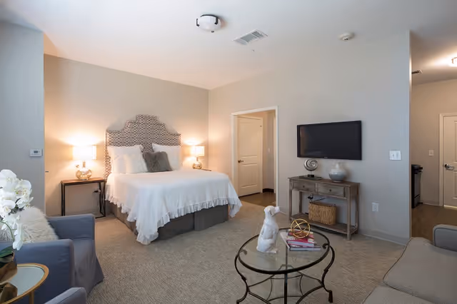 A cozy senior living bedroom with a queen-sized bed featuring a patterned headboard and white bedding. Two bedside tables with lamps flank the bed. There is a gray armchair with a white pillow and a small round glass coffee table with decorative items in front of a gray sofa. A flat-screen TV is mounted on the wall above a wooden console table with decorative objects. The room has beige walls and carpeted flooring, with two doorways visible leading to other rooms.