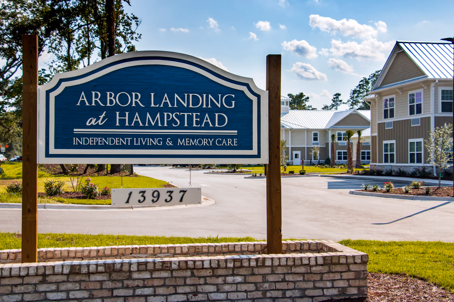 A large blue and white sign for Arbor Landing at Hampstead, an independent living and memory care facility, is displayed in front of a paved driveway and landscaped grounds with buildings in the background under a partly cloudy sky.