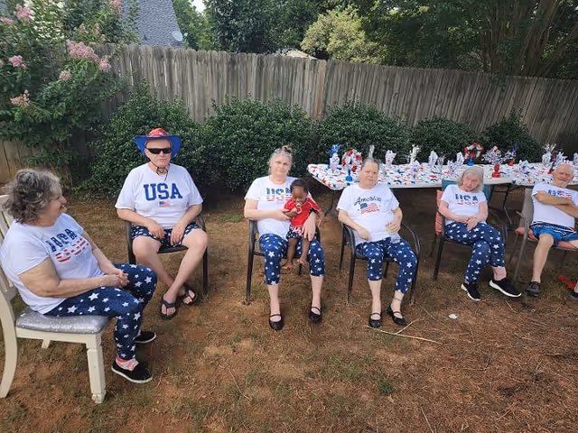 A group of six elderly people sitting outdoors on chairs in a garden area with a wooden fence and bushes in the background. They are dressed in patriotic clothing featuring USA-themed shirts and star-spangled pants. One person is holding a small child dressed in red. A table behind them is decorated with red, white, and blue items.