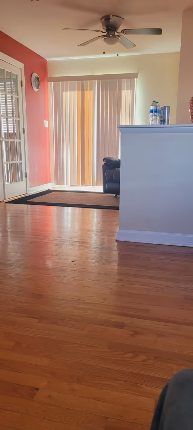 Interior view of a living room area with wooden flooring, a ceiling fan, vertical blinds covering a sliding glass door, a black leather armchair partially visible behind a white half-wall, and a red accent wall with a clock and a glass-paneled door.