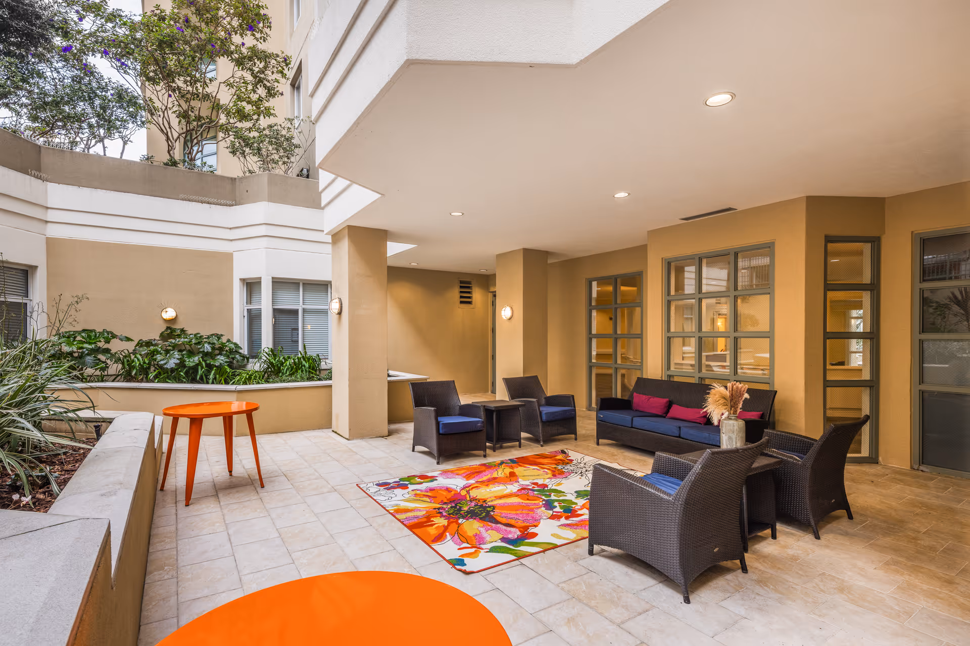 Covered outdoor seating area with wicker chairs and a sofa with blue cushions and pink pillows, a colorful floral rug on the tiled floor, orange tables, and plants along the walls under a beige building structure.