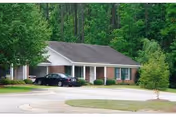 Single-story residential building with a gray roof and white columns on the porch, surrounded by green trees and a curved driveway with a black car parked in front.