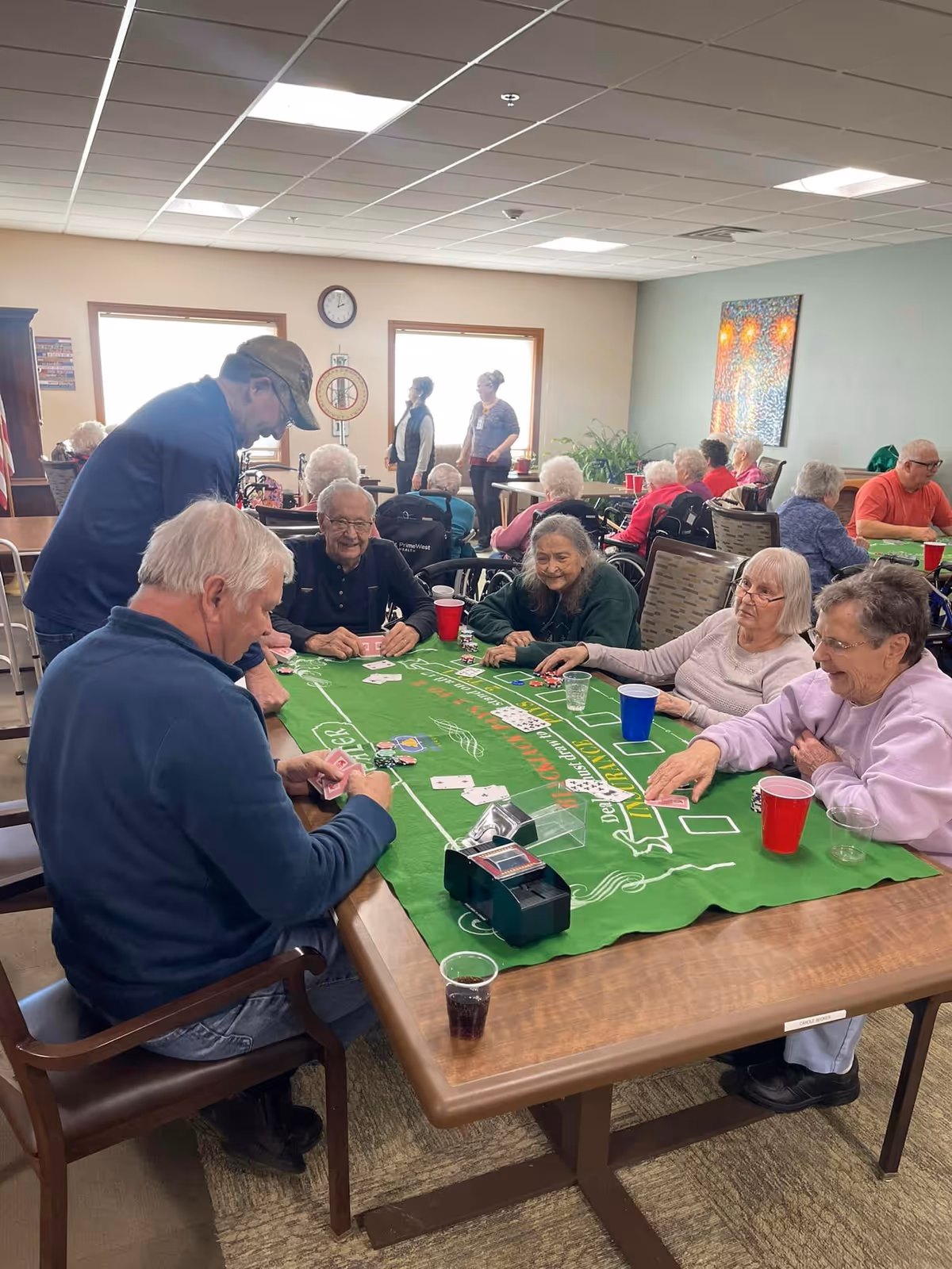 A group of elderly people sitting around a table playing a card game on a green felt table cover in a well-lit common room. Several other elderly individuals are seated or standing in the background, engaging in various activities. The room has large windows, a clock on the wall, and colorful artwork. Red and blue plastic cups are on the table.