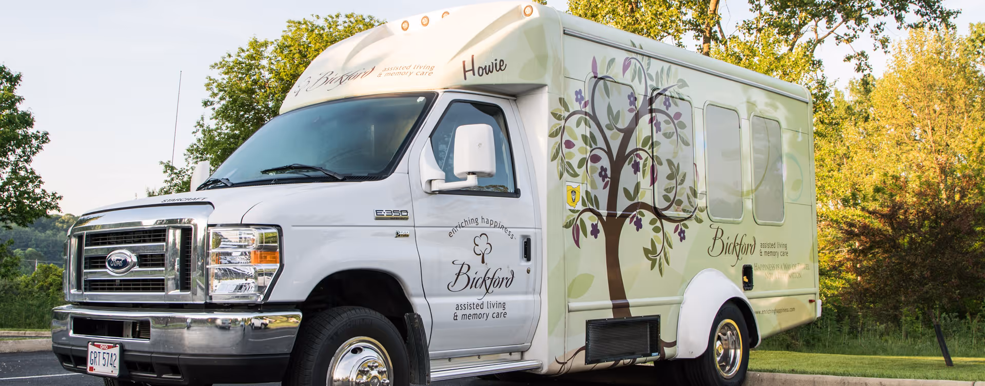 A white and light green shuttle bus with floral and tree designs parked outdoors near trees and grass. The bus has the name 'Bickford' and the phrases 'assisted living & memory care' and 'enriching happiness' written on its side. The front license plate reads 'Ohio GRT 5742'.