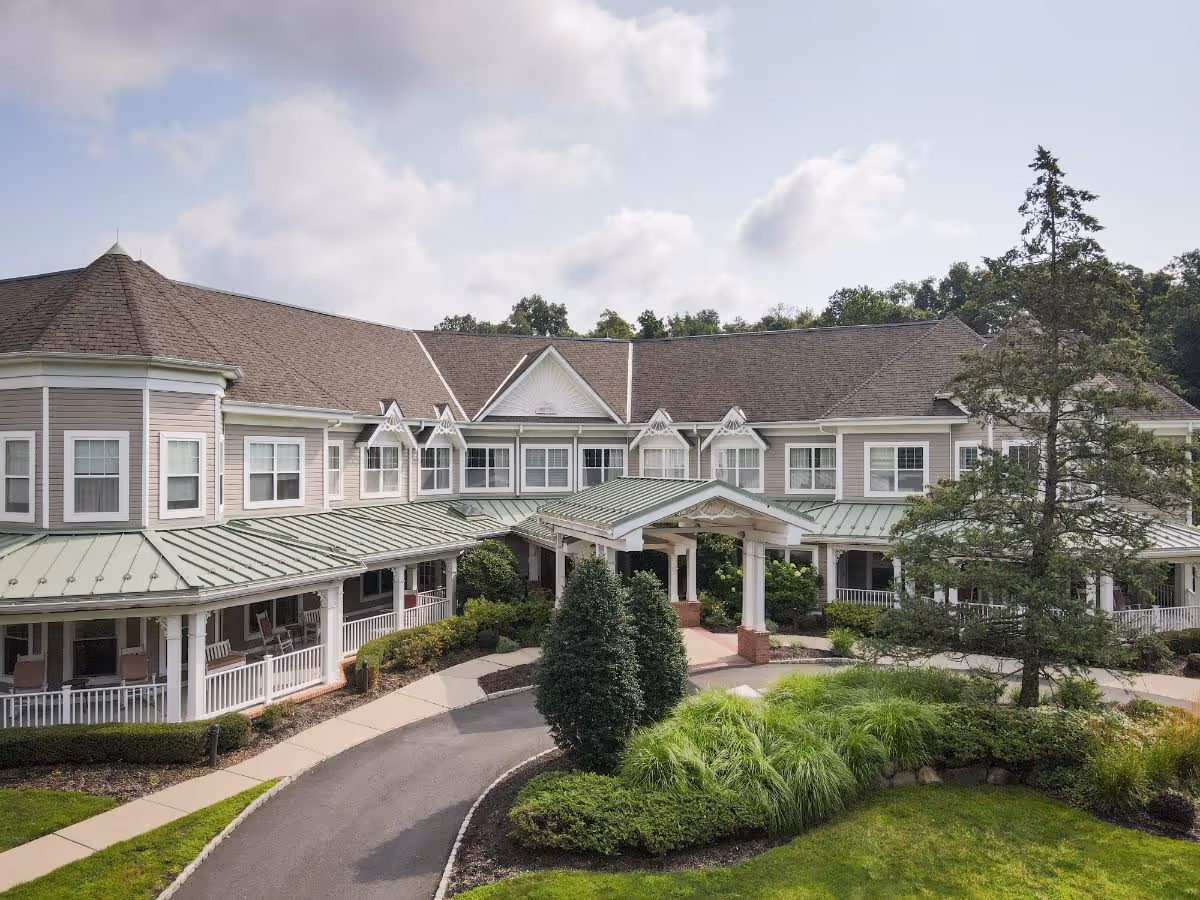 Front exterior of a two-story senior living building with a covered entrance, wraparound porch, and landscaped circular driveway.