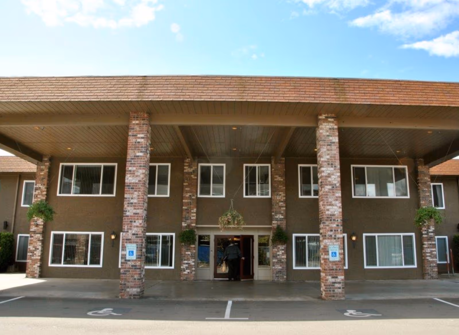 Front exterior view of a two-story building with a covered entrance supported by brick columns. There are several windows with white frames and hanging plants near the entrance. The sky is clear with a few clouds, and there are two handicap parking spaces visible in front of the building. A person is entering through the main doors.