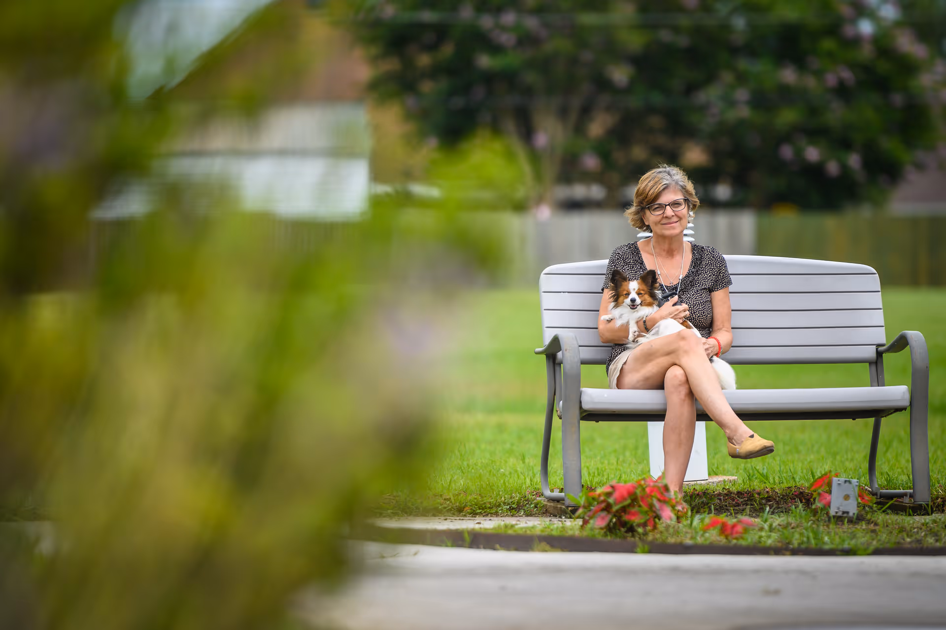A woman sitting on a gray bench outdoors, holding a small dog in her lap, with green grass and trees in the background.
