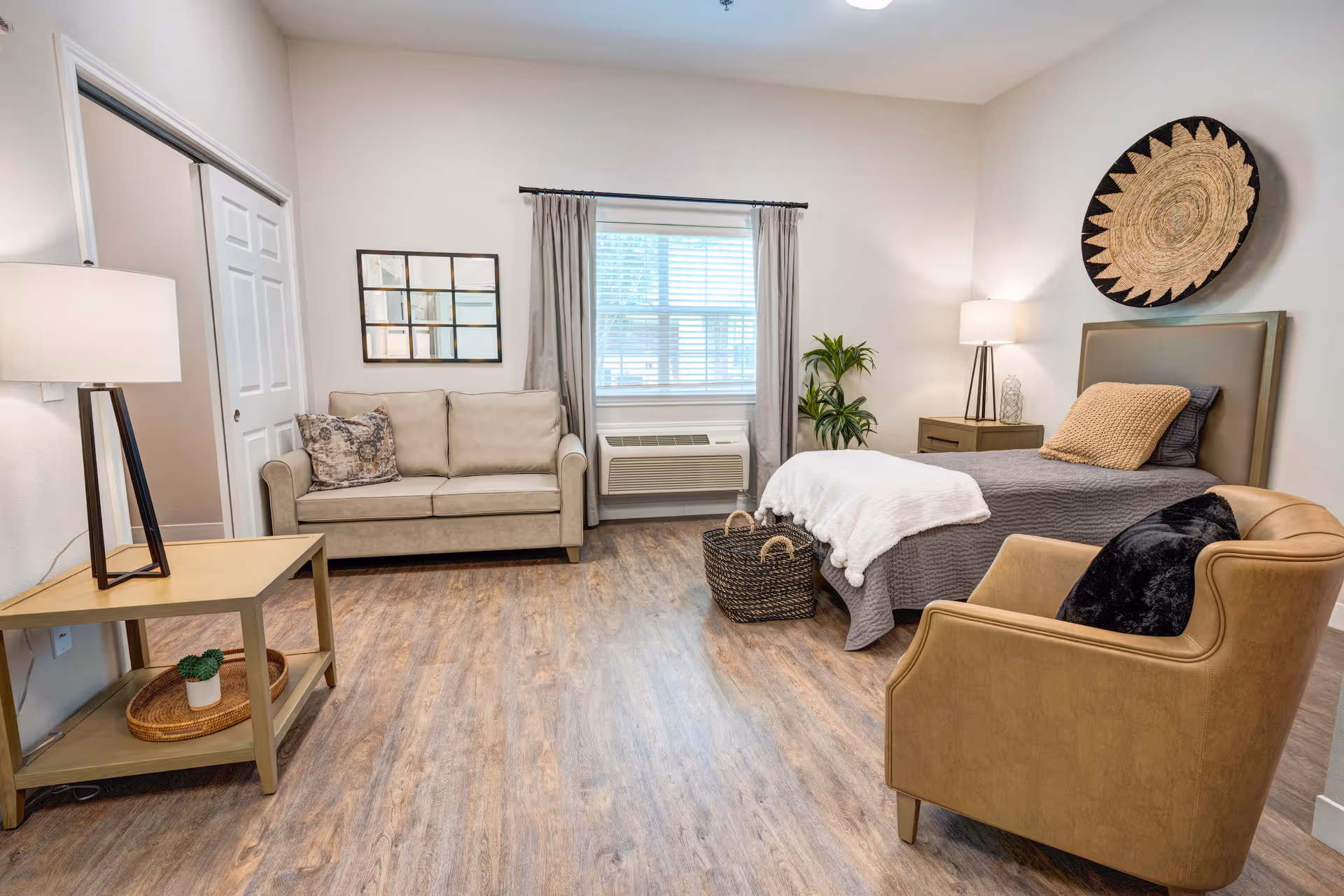 A cozy and well-lit assisted living bedroom featuring a single bed with gray bedding and a beige throw pillow, a tan armchair with a black pillow, a beige loveseat with a decorative pillow, a wooden side table with a lamp and a small plant, a window with gray curtains, a woven basket on the floor, and a large decorative wall piece above the bed.