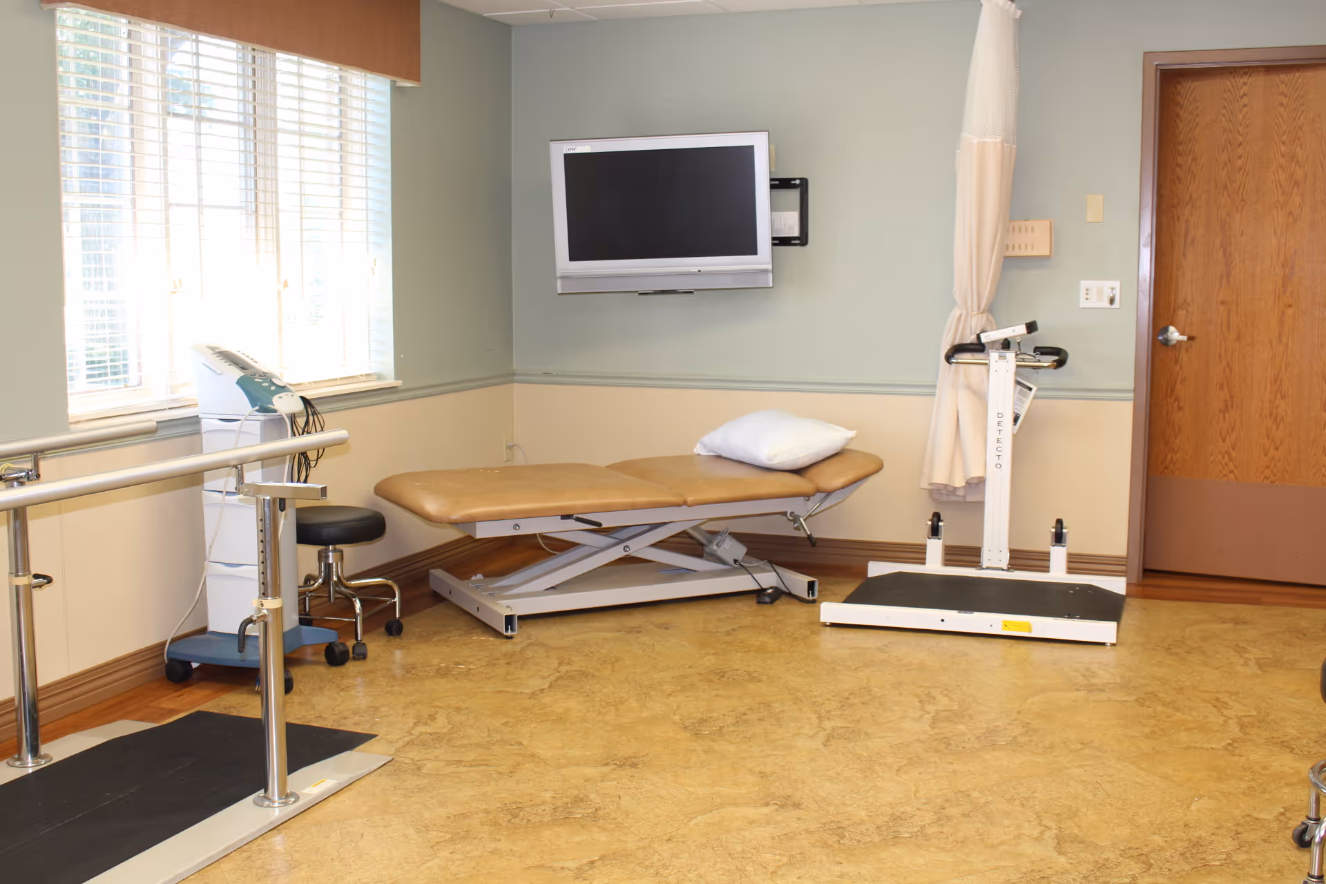 A therapy or rehabilitation room with a padded adjustable treatment table, a pillow, a wall-mounted flat screen TV, parallel bars for walking exercises, a stool, a medical device on a stand, and a scale. The room has a window with blinds and a wooden door.