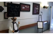 A hallway in a nursing and rehab center with a mannequin dressed as a chef holding a chalkboard sign that reads 'Main Street Cafe - Now Open'. The hallway has framed pictures on the wall, a wall-mounted light fixture, and an easel with a blank sign near a door.