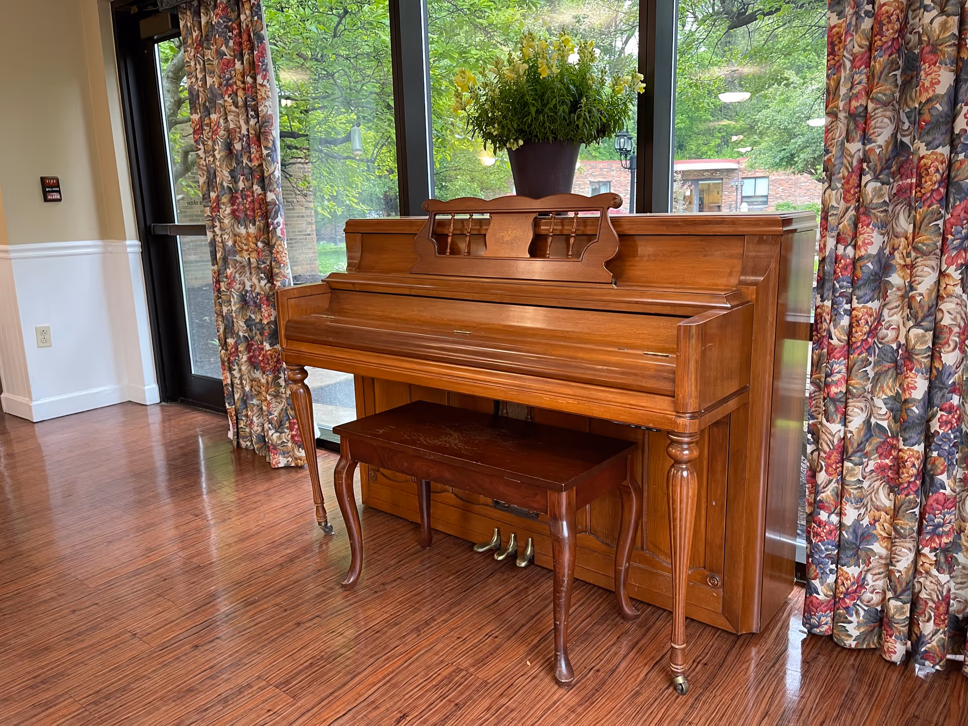 A wooden upright piano and bench sit in a lobby area in front of glass doors with floral curtains.