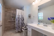Accessible bathroom with a walk-in tiled shower and shower chair, white vanity with sink and mirror, and a window with light coming in.