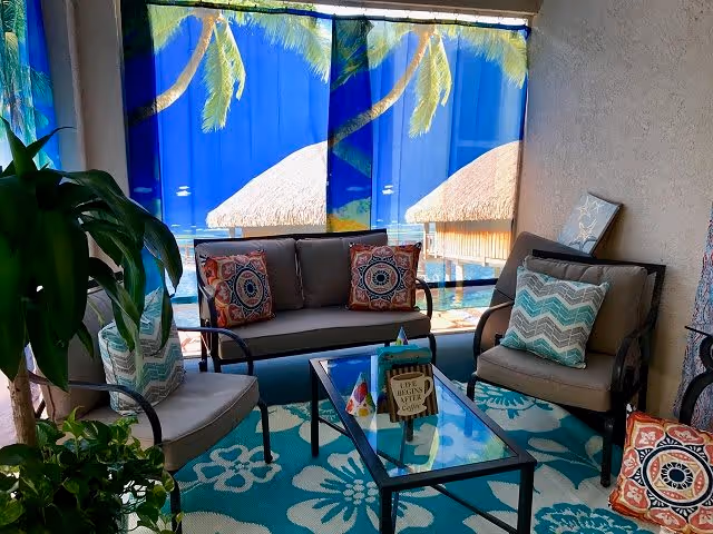 A cozy indoor seating area with a sofa and two armchairs arranged around a glass coffee table. The furniture has patterned cushions in various colors. The floor has a blue and white floral rug. Behind the seating area, there are large windows covered with curtains depicting a tropical beach scene with palm trees and thatched huts over water. There are also some green plants visible on the left side.