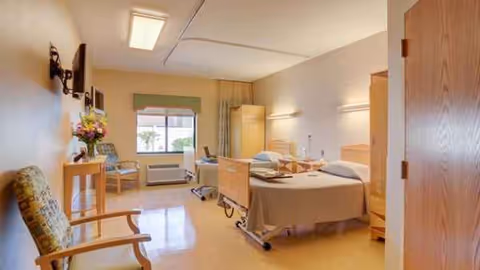 Two hospital-style beds in a bright senior living room with chairs and a window.