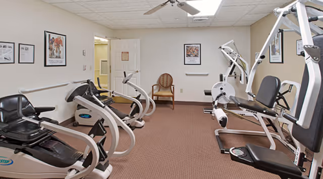 A small fitness room with exercise equipment including recumbent bikes and a multi-function weight machine. The room has beige walls with framed motivational posters, a ceiling fan, and a single wooden chair against the far wall. The floor is carpeted in brown.