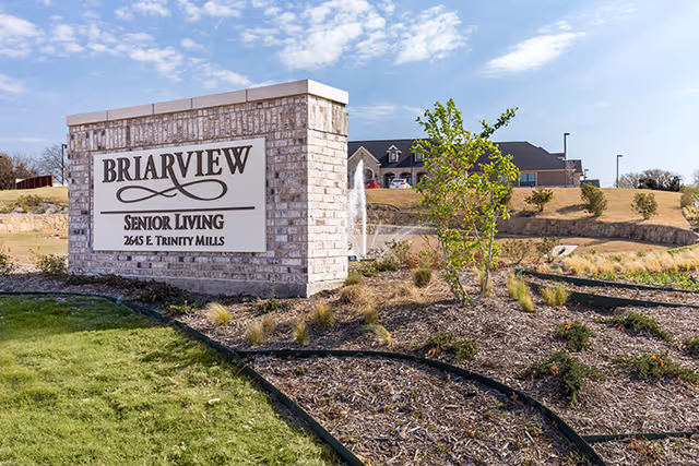 A large brick sign for Briarview Senior Living at 2645 E. Trinity Mills is displayed outdoors with a landscaped area of grass, mulch, and small plants around it. In the background, there is a building and a water fountain under a partly cloudy sky.