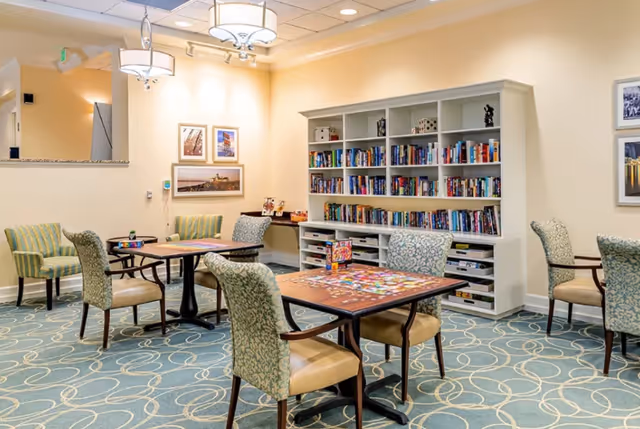 Bright communal activity room with tables and upholstered chairs, a large bookshelf of books, and puzzles on the tabletops.