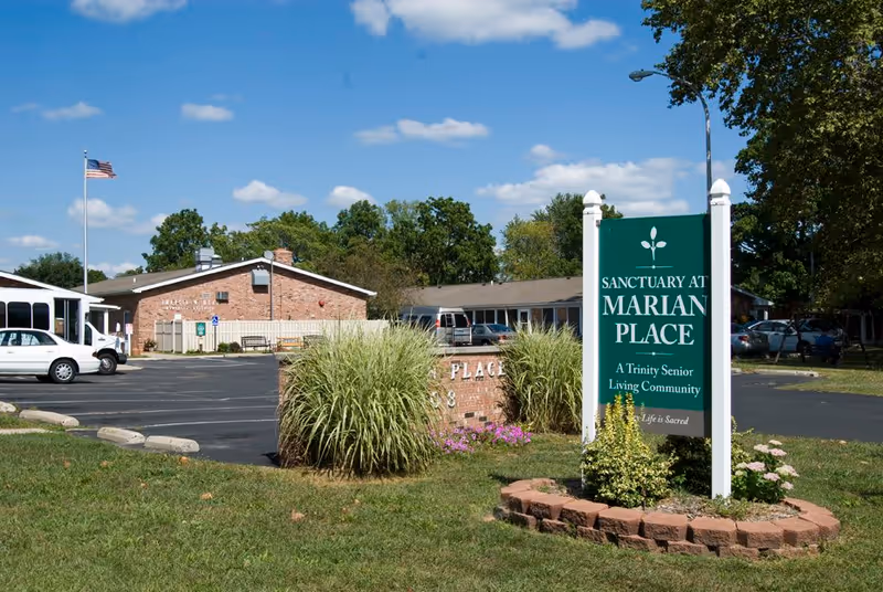 Exterior view of Sanctuary at Marian Place senior living community showing a parking lot, a brick building, an American flag on a pole, and a green sign with white text surrounded by plants and grass under a blue sky with scattered clouds.