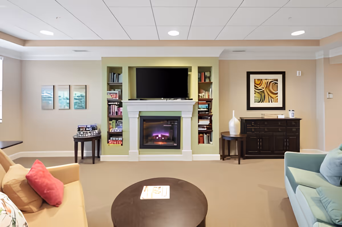 A bright communal living room with sofas facing a central fireplace topped by a wall-mounted TV and flanked by built-in shelves and side tables.