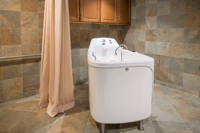 A white walk-in bathtub in a tiled room with a beige shower curtain and wooden cabinets overhead.