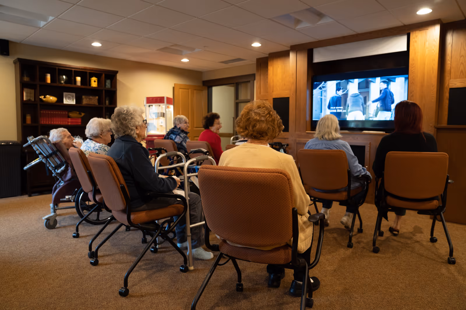A group of elderly individuals seated in chairs and wheelchairs watching a television in a cozy room with wood-paneled walls and a bookshelf in the background.