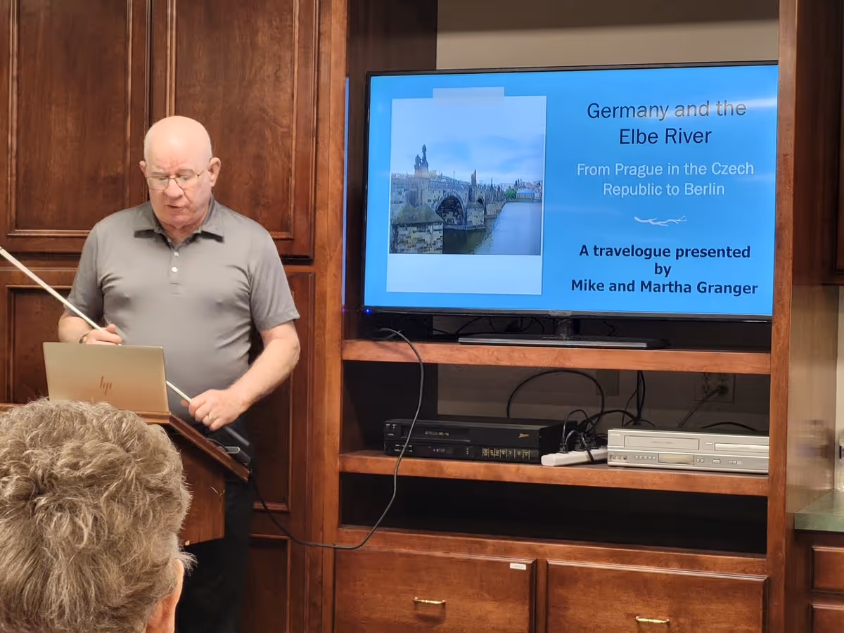 An elderly man wearing a gray polo shirt is giving a presentation in a room with wooden cabinets. He is standing behind a podium with a laptop and pointing at a screen displaying a slide titled 'Germany and the Elbe River From Prague in the Czech Republic to Berlin A travelogue presented by Mike and Martha Granger' with an image of a bridge over a river.