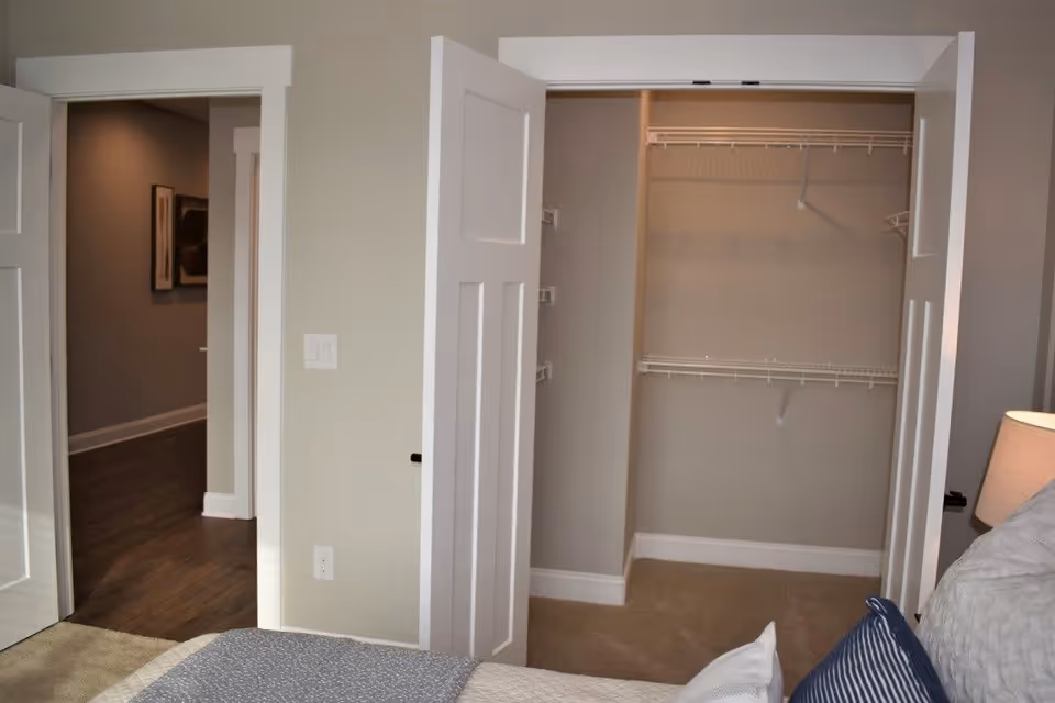 Bedroom interior showing an open closet with wire shelving, part of a bed, and a doorway to a hallway.