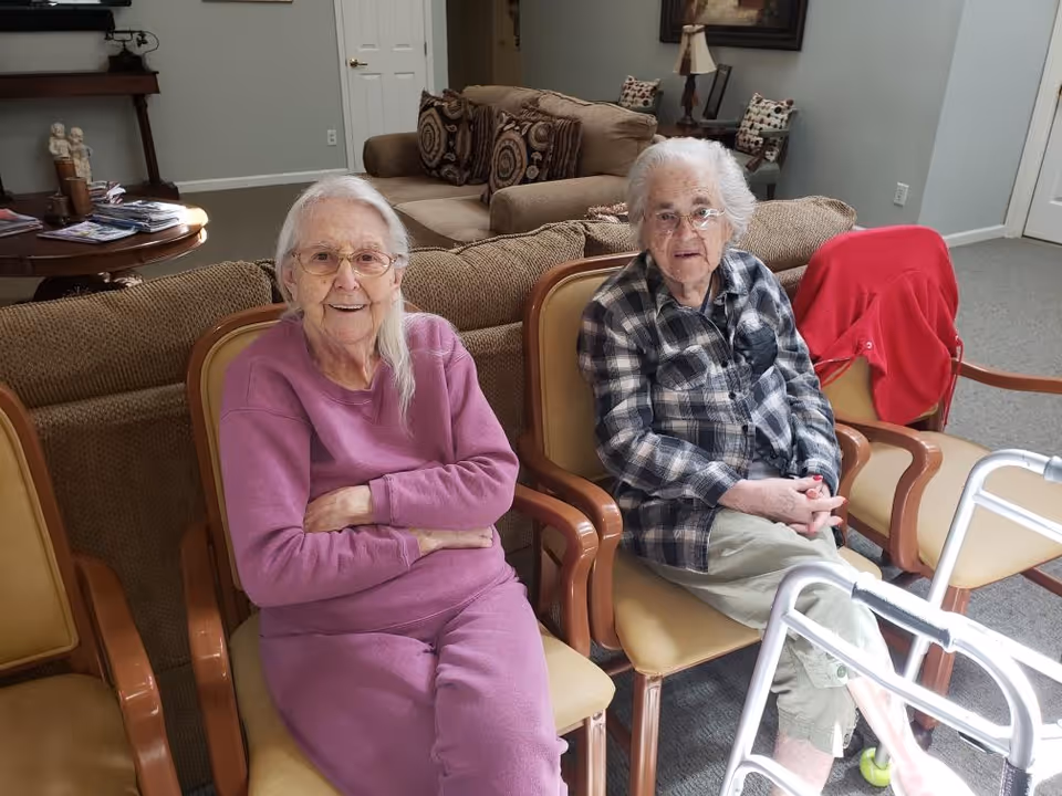 Two elderly women sitting on chairs in a living room area of an assisted living facility. One woman is wearing a purple outfit and glasses, smiling with her arms crossed. The other woman is wearing a plaid shirt and light-colored pants, sitting next to a walker. Behind them is a couch with patterned pillows and a table with magazines and decorative items.