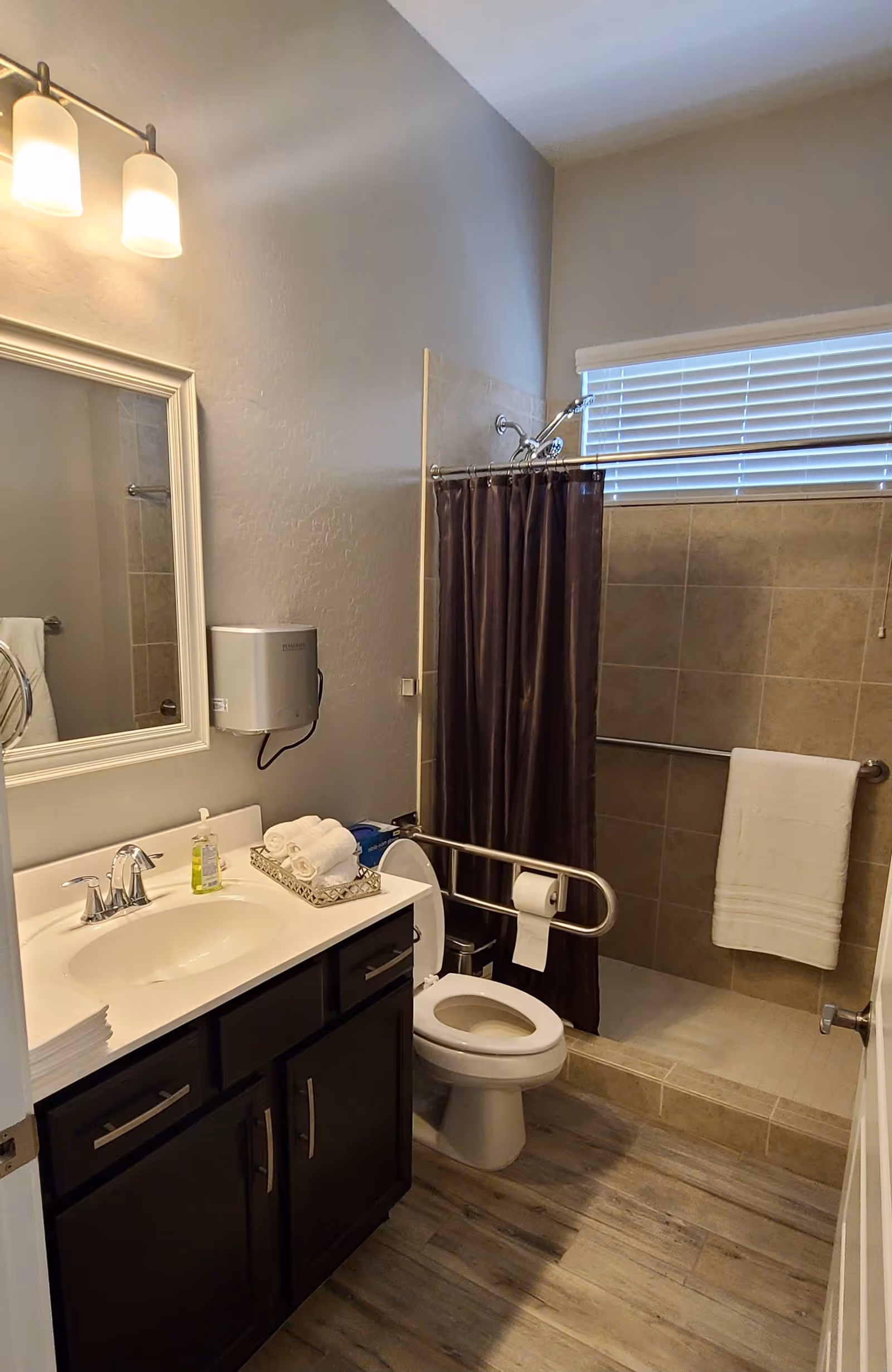 A clean and accessible bathroom with a white sink and dark wood cabinet, a large mirror with two light fixtures above it, a toilet with a safety grab bar and toilet paper holder, and a walk-in shower with a dark shower curtain and a towel hanging on a grab bar. The floor has wood-like tiles and the walls are painted light gray.