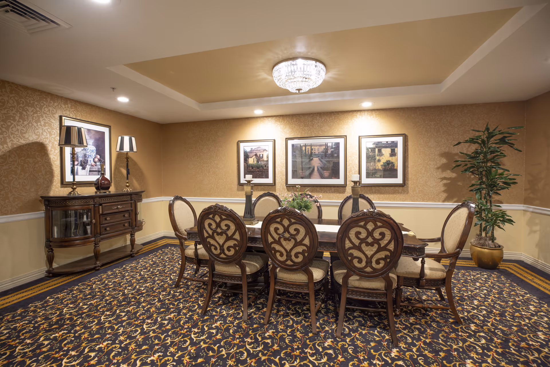 A formal dining room with an ornate wooden dining table surrounded by eight upholstered chairs. The room features patterned carpet, beige wallpaper with a decorative border, three framed paintings on the wall, a sideboard with two lamps and a vase, a potted plant in the corner, and a crystal chandelier on the ceiling.