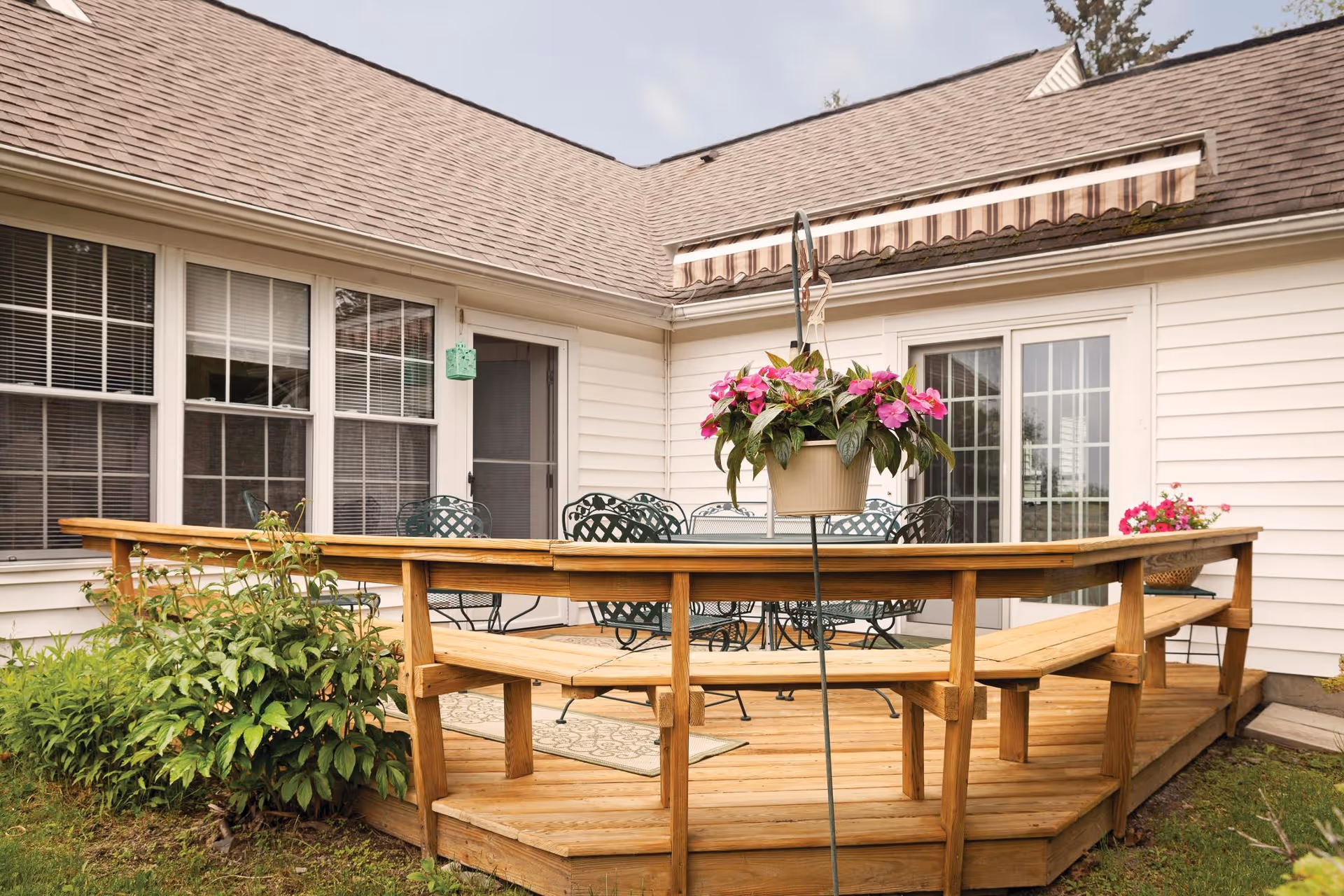 Outdoor wooden deck attached to a white house with a set of metal chairs and a table. The deck has built-in wooden benches and a hanging flower pot with pink flowers. There are windows and a glass door leading into the house, with an awning above the door.