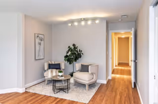 A cozy sitting area in a senior living facility with two beige armchairs, a small round coffee table with a plant, a large leafy plant in the corner, a framed artwork on the wall, and a hallway leading to other rooms. The floor is wooden and there is a light fixture on the ceiling.