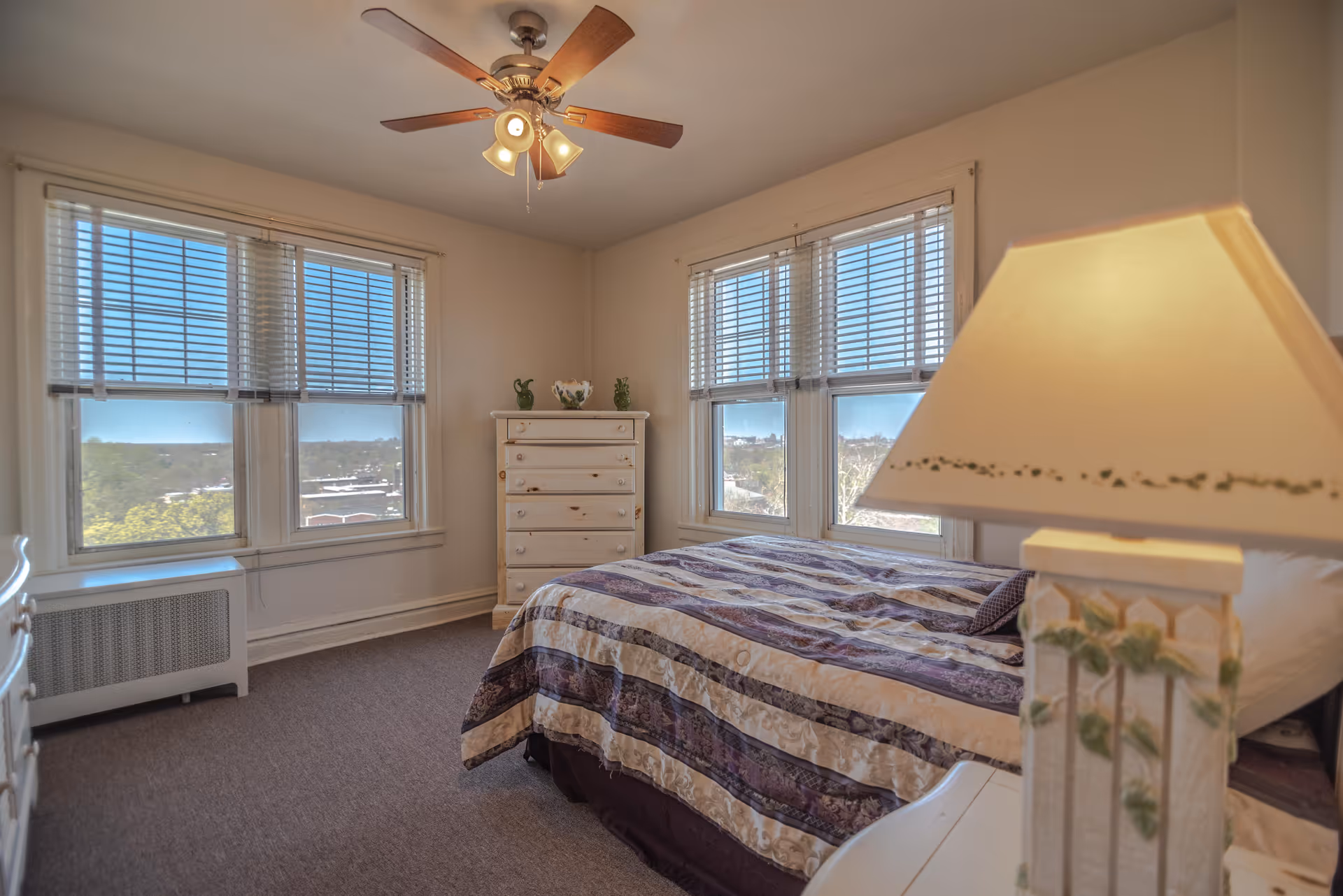 A bright bedroom with a bed covered in a purple and beige striped comforter, a white wooden dresser with decorative items on top, large windows with blinds letting in natural light, a ceiling fan with lights, and a bedside lamp with a floral design.