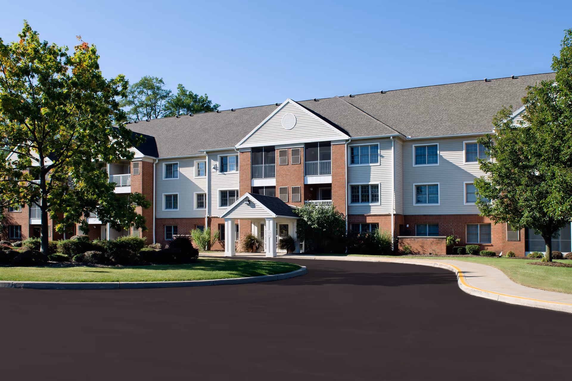 Exterior view of a three-story senior living facility building with a combination of brick and light-colored siding, surrounded by green trees and landscaping under a clear blue sky. A curved driveway leads to the entrance with a small covered porch.