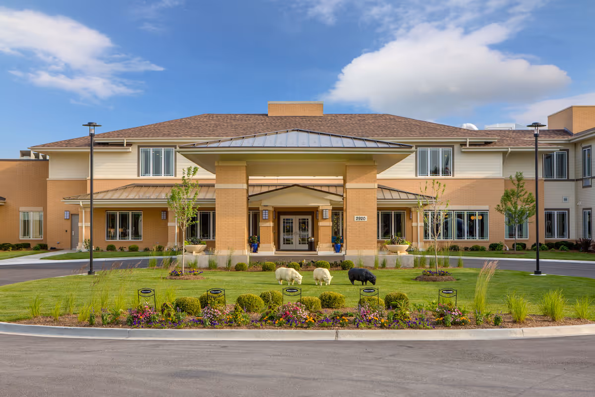 Front exterior view of Arbor Terrace Naperville, a two-story senior living facility with a covered entrance, manicured lawn, flower beds, and decorative sheep statues on the grass under a partly cloudy sky.