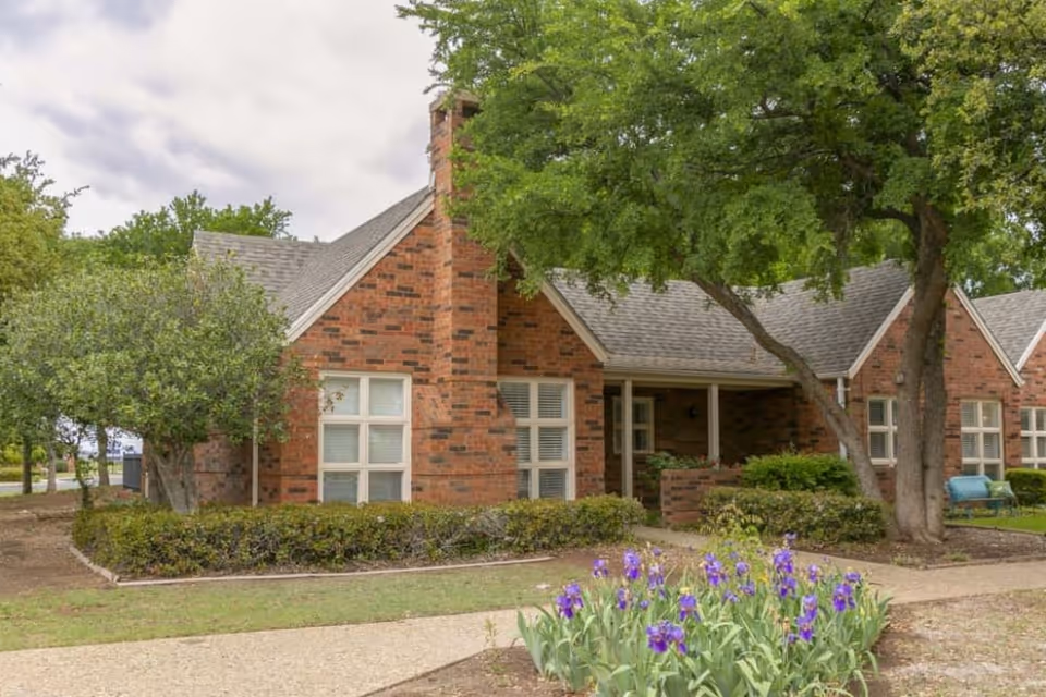 Exterior view of a brick building with multiple windows and a chimney, surrounded by trees, bushes, and a flower bed with purple flowers in front. A paved walkway curves through the landscaped yard.