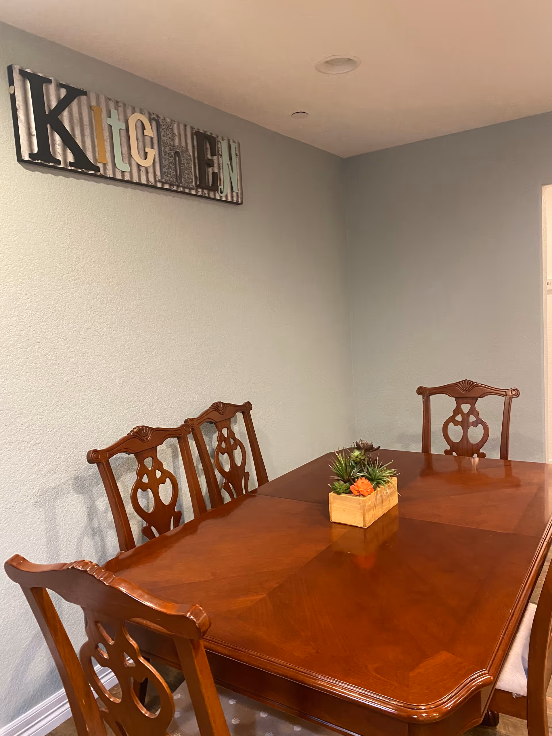 A wooden dining table with four matching chairs in a room with light gray walls. A small rectangular planter with succulents is centered on the table. On the wall above the table is a decorative sign spelling out 'KITCHEN' with each letter in a different style and color.