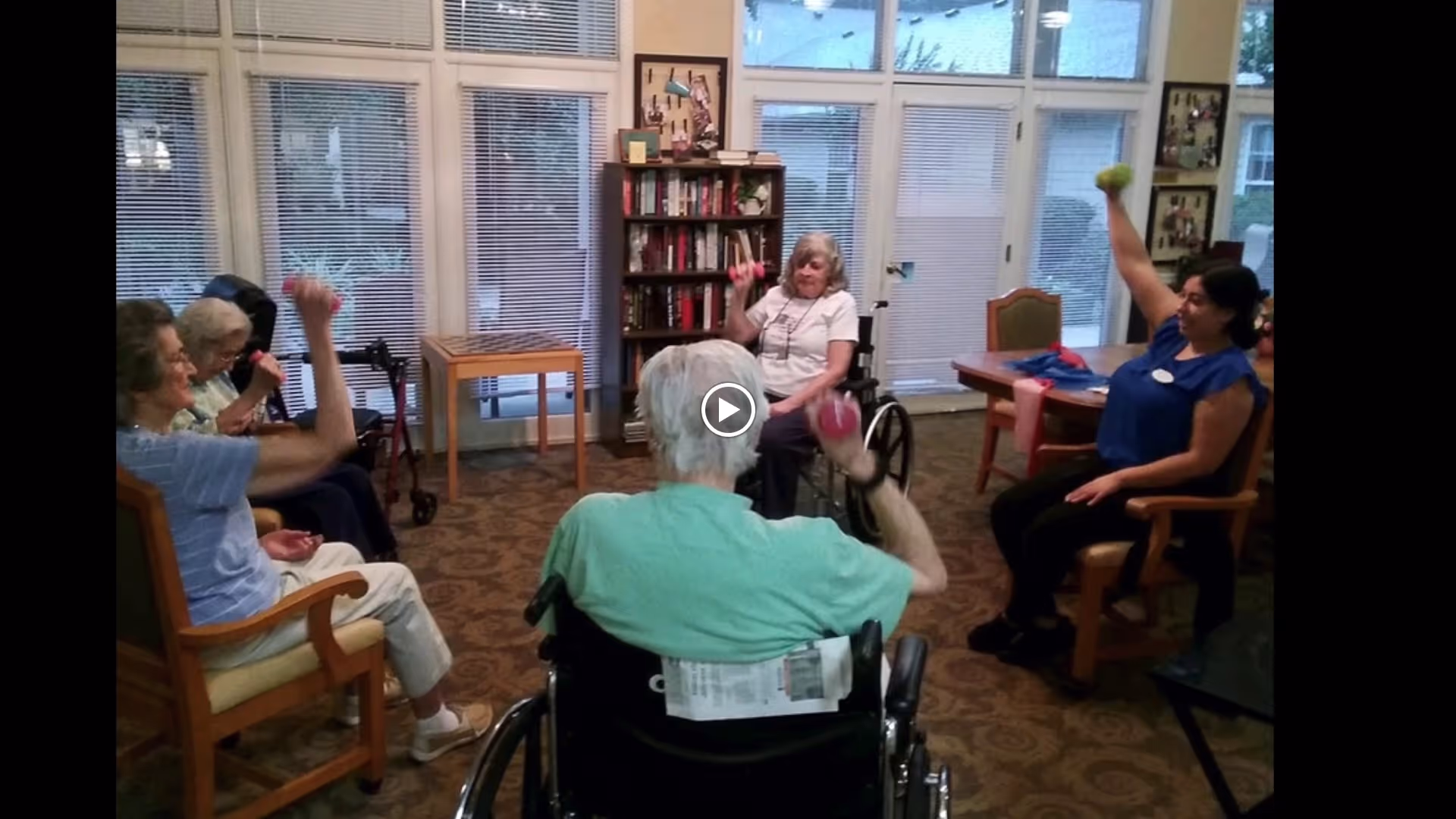 Residents and a staff member seated in a communal room doing seated arm exercises with small hand weights.