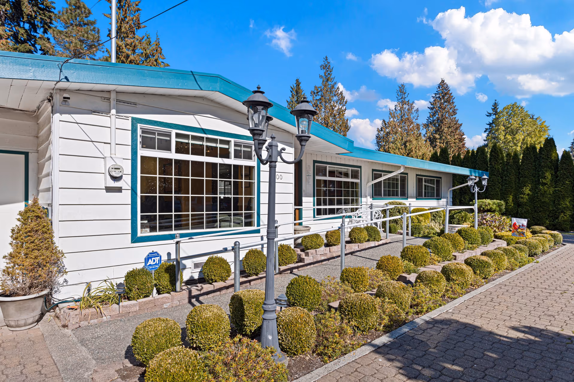 Exterior view of a single-story senior care facility building with white siding and teal trim. The building has large windows and a ramp with handrails leading to the entrance. There are neatly trimmed round bushes and a paved walkway in front, along with two black vintage-style street lamps. Tall evergreen trees and a bright blue sky with some clouds are visible in the background.