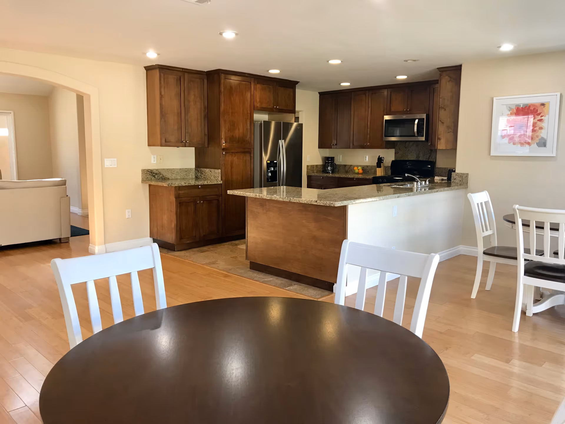 Interior view of a senior living facility kitchen and dining area. The kitchen features wooden cabinets, granite countertops, a stainless steel refrigerator, microwave, and a stove. In the foreground, there is a round dark wooden dining table with white chairs. The room has light-colored walls, recessed ceiling lights, and a framed floral artwork on the wall.