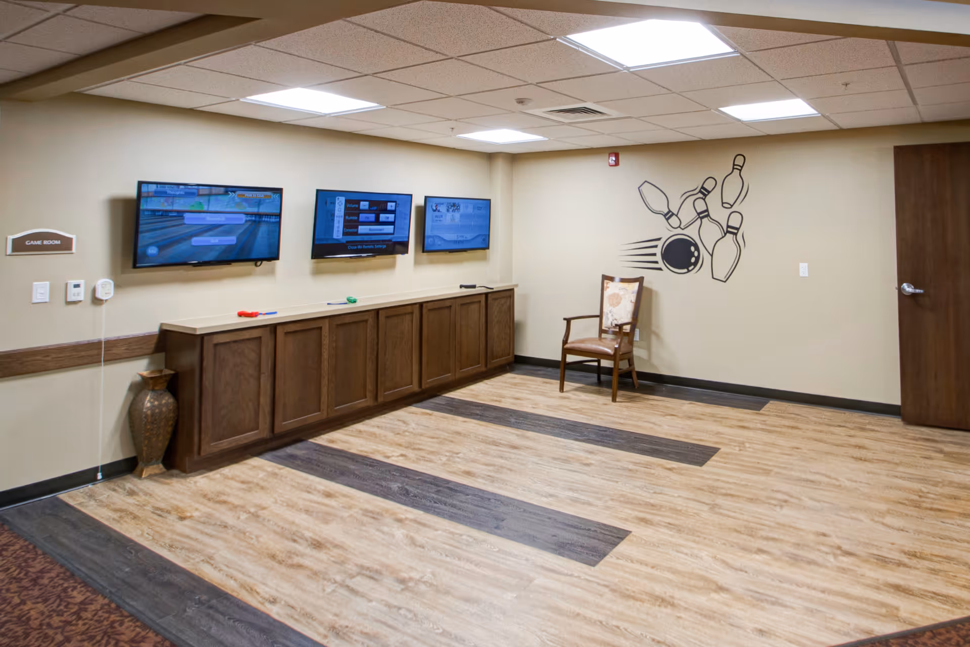 A game room with three wall-mounted flat screen TVs displaying a bowling game. Below the TVs is a long wooden cabinet. There is a single wooden chair with a cushioned seat and backrest against the wall. The wall features a decorative graphic of a bowling ball hitting pins. The floor has a wood pattern with darker wood strips, and the ceiling has recessed lighting.