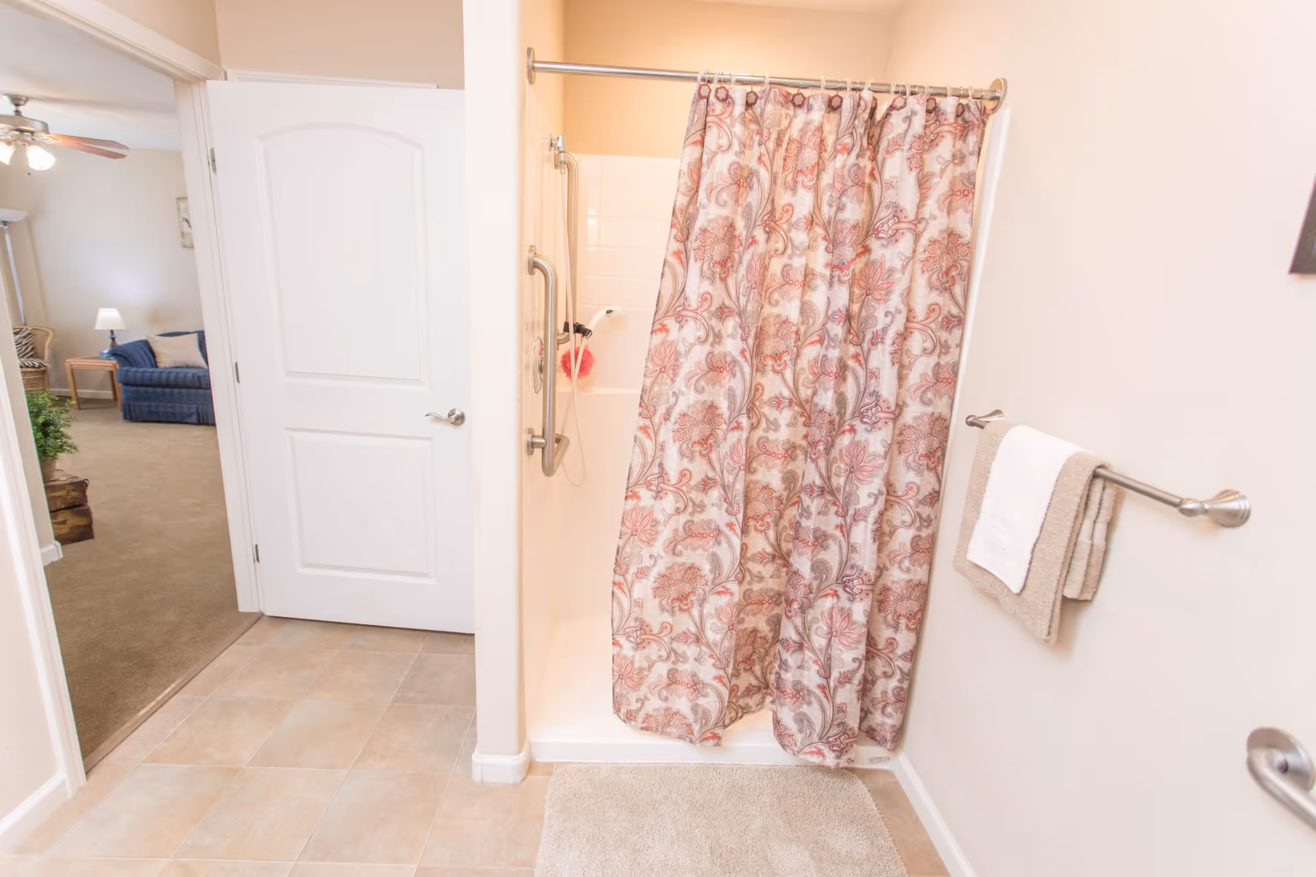 Bathroom with a shower area featuring a floral patterned shower curtain, grab bars, and a handheld showerhead. A towel rack with beige and white towels is mounted on the wall. The bathroom floor is tiled, and an open door leads to a carpeted living room area with a blue couch, side table, lamp, and ceiling fan.