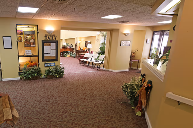 Interior view of a senior living facility hallway with carpeted floor and beige walls. There is a bulletin board labeled 'Brookdale Daily' on the left wall, some plants on the floor, and a few white folding chairs arranged in the background near a seating area with more plants and furniture. The space is well-lit with ceiling lights and wall sconces.