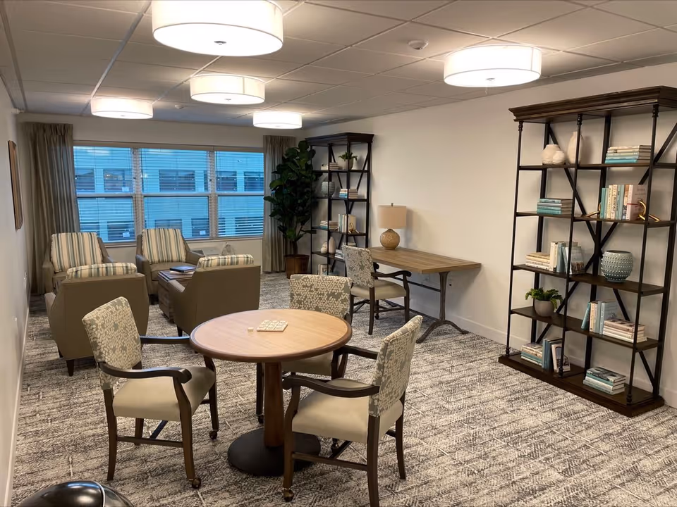 A cozy common area in a senior living facility featuring a round wooden table with four upholstered chairs, two armchairs with striped cushions near a large window, two tall bookshelves filled with books and decorative items, a wooden desk with a lamp, and soft overhead lighting.