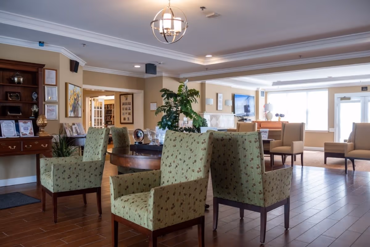 A spacious and well-lit common area in a senior living facility with several upholstered armchairs arranged around a round wooden table. The room features wooden flooring, beige walls, a chandelier, and a large window letting in natural light. There is a wooden cabinet with framed certificates and decorative items on the left, a plant on the table, and a flat-screen TV mounted on the far wall.