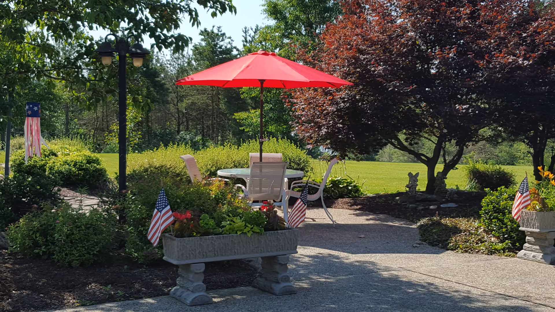 Outdoor garden area with a round glass table and four chairs under a red umbrella. The area is surrounded by green bushes, trees, and flower planters decorated with small American flags. A lamp post and a decorative wind sock are visible in the background.